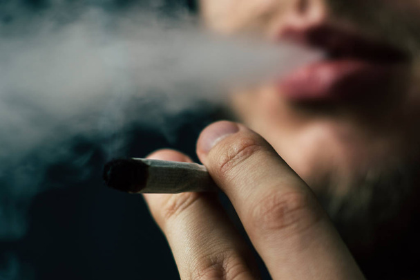 Rauch auf schwarzem Hintergrund. Ein Mann raucht Cannabiskraut, einen Joint und ein Feuerzeug in den Händen. Konzepte des medizinischen Marihuana-Konsums und der Legalisierung von Cannabis. - Foto, Bild