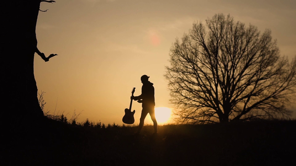 Sziluettjét gitárosa naplemente lassított. Gitáros sétál egy gitárral a kezében - Felvétel, videó