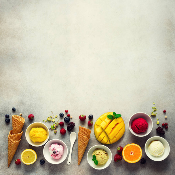 Top view red, pink, yellow, green, white ice cream balls in bowls, waffle cones, berries, orange, mango, pistachio, grey concrete background. Colorful collection, flat lay, summer concept - Foto, Bild