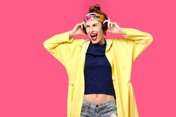 Nuori kaunis nainen vaaleanpunaisella taustalla ja keltainen takki kuuntelemassa musiikkia langattomien kuulokkeiden kautta. Terveellisen ravitsemuksen ja urheilun käsite. Väripakkomielle. Minimalistinen tyyli. Pietari
 - Valokuva, kuva