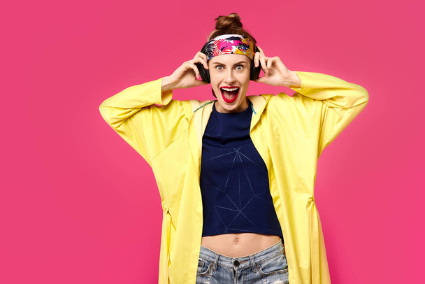 Nuori kaunis nainen vaaleanpunaisella taustalla ja keltainen takki kuuntelemassa musiikkia langattomien kuulokkeiden kautta. Terveellisen ravitsemuksen ja urheilun käsite. Väripakkomielle. Minimalistinen tyyli. Pietari
 - Valokuva, kuva