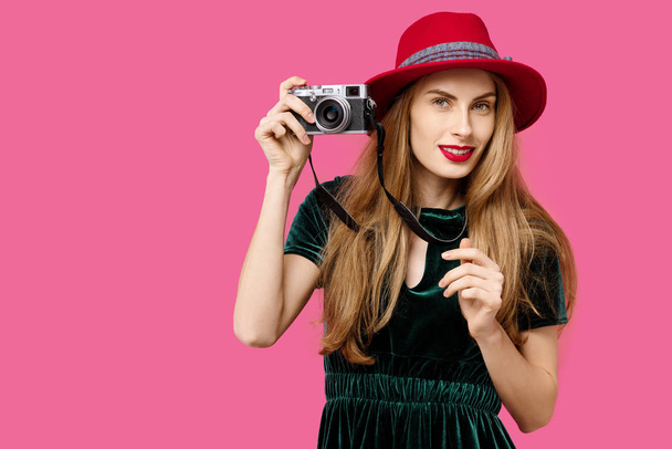 junge schöne Frau auf rosa Hintergrund in grünem Kleid und Hut hält eine Seltenheitskamera in den Händen und lächelt. Konzept der gesunden Ernährung und Sport. Farbbesessenheit. Minimalistischer Ansatz - Foto, Bild