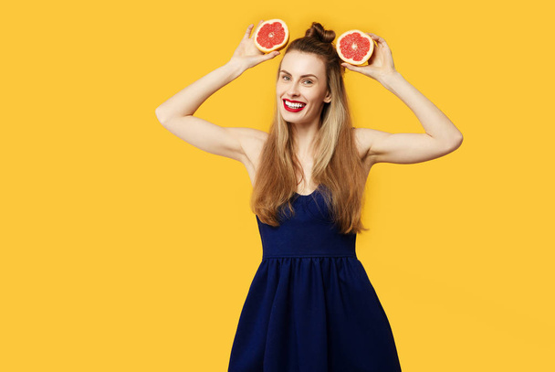Молодая женщина на жёлтом фоне и в синем платье держит порезанный оранжевый в руках. Концепция здорового питания и спорта. Концепция цветовой одержимости. Минималистический стиль. Стильный тренд
 - Фото, изображение
