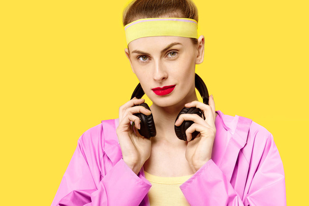 junge Frau auf gelbem Hintergrund und rosa Mantel umarmt Musikwurm drahtlose Kopfhörer. Farbbesessenheit. Minimalistischer Stil. modisch trendy - Foto, Bild
