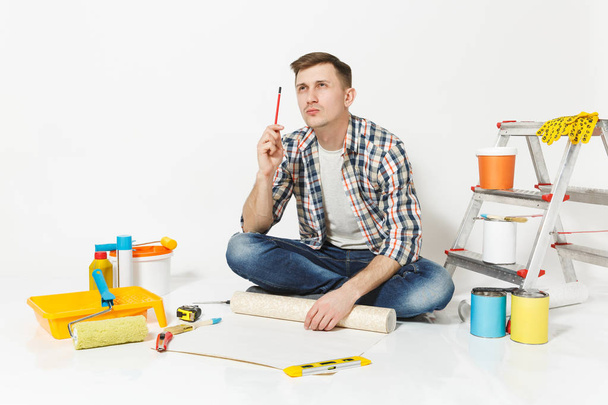 Young man sitting on floor with wallpaper roll, pencil, instruments for renovation apartment room isolated on white background. Wallpaper, gluing accessories, painting tools. Repair home concept. - Photo, Image