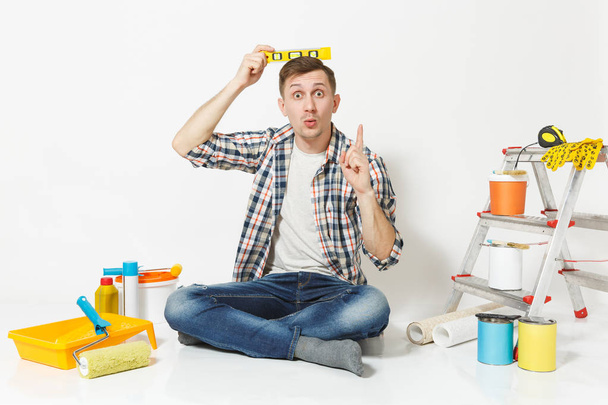 Διασκέδαση ευτυχής άνδρας κάθεται στο πάτωμα με την οικοδόμηση αλφάδι φυσαλίδας στο κεφάλι, όργανα για διαμέρισμα ανακαίνιση που απομονώνονται σε λευκό φόντο. Ταπετσαρία, κόλλημα εξαρτήματα, εργαλεία. Επισκευή concept home. - Φωτογραφία, εικόνα