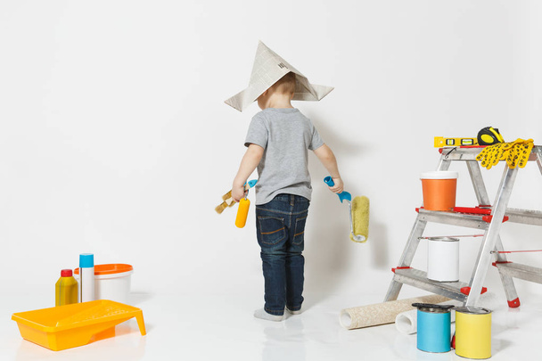 Little cute boy in newspaper hat with instruments for renovation apartment room isolated on white background. Wallpaper, gluing accessories, painting tools. Repair home. Parenthood, childhood concept. - Photo, Image