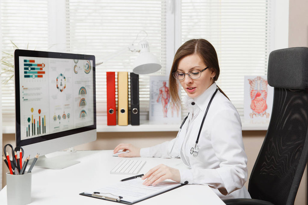 Young woman sitting at desk, working on computer, filling out medical documents in light office in hospital. Female doctor in medical gown, stethoscope in consulting room. Healthcare, medicine concept - Photo, Image