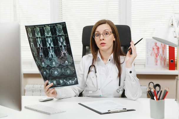 Женщина-врач, сидящая за столом с компьютером, делает рентген мозга с помощью рентгеновского снимка КТ в легком кабинете больницы. Женщина в медицинском халате в кабинете консультанта. Здравоохранение, медицина
. - Фото, изображение