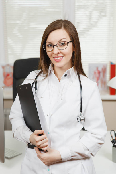 Young smiling female doctor standing at desk with clipboard and medical documents in light office in hospital. Woman in medical gown, stethoscope in consulting room. Healthcare, medicine concept. - Zdjęcie, obraz