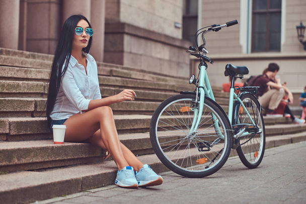 Een sexy brunette vrouw dragen van blouse en denim shorts in zonnebril, ontspannen na het rijden op een fiets, zitten met een kopje koffie op stappen in een stad. - Foto, afbeelding