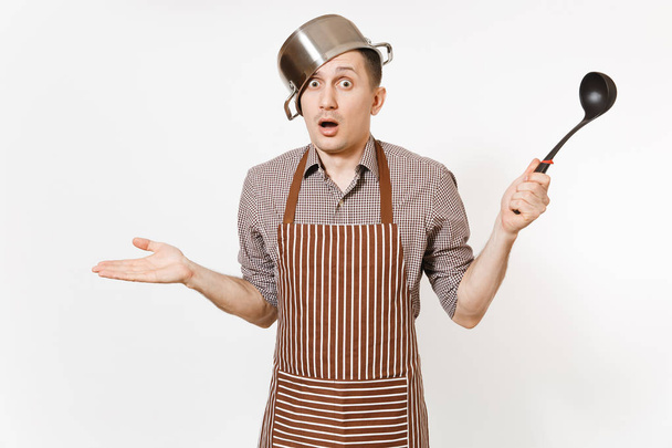 Fun mad man in striped apron with ladle, silver stainless glossy aluminium empty stewpan, pan or pot on head isolated on white background. Male housekeeper or houseworker. Kitchenware, dishes concept. - Photo, image