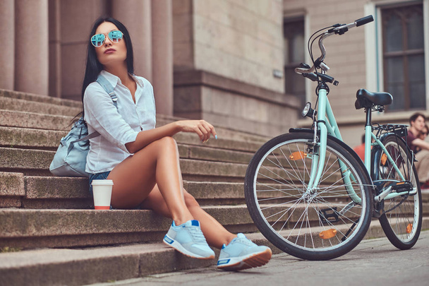 Uma mulher morena sexy vestindo blusa e calções jeans em óculos de sol, relaxando depois de andar de bicicleta, sentada com uma xícara de café em passos em uma cidade
. - Foto, Imagem