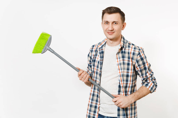 Young smiling happy housekeeper man in checkered shirt holding and sweeping with green broom isolated on white background. Male doing house chores. Copy space for advertisement. Cleanliness concept. - Foto, Imagem