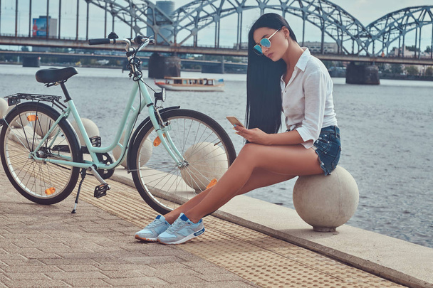Porträt eines sexy brünetten Mädchens in Bluse und Shorts mit Sonnenbrille, das auf einem Kai an der Brücke sitzt, ein Smartphone benutzt und sich nach dem Fahrradfahren entspannen kann. - Foto, Bild