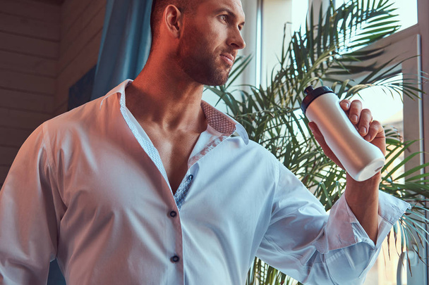 Close-up πορτρέτο του ένα ψηλός γενειοφόρος άνδρας με ένα μυώδες σώμα, ντυμένος με ένα άσπρο πουκάμισο, πρωτεΐνη ποτό πριν από μια προπόνηση το πρωί. - Φωτογραφία, εικόνα