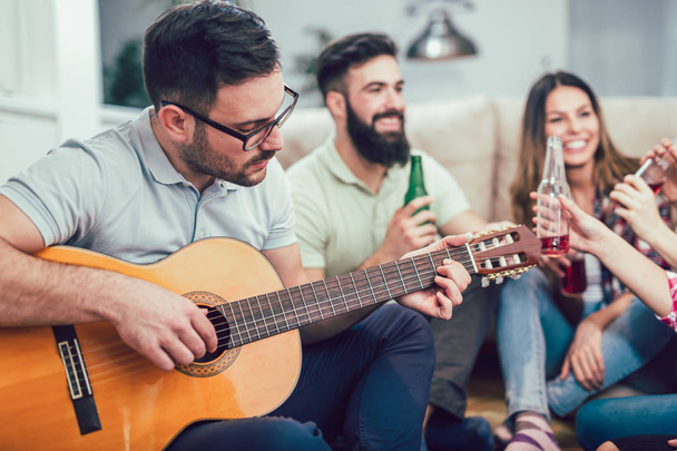 Χαρούμενος νεαρός παρέα με κιθάρα διασκεδάζοντας και πίνοντας μπύρα στο σπίτι εσωτερικό - Φωτογραφία, εικόνα