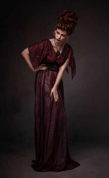Full length view of woman with baroque hairstyle and evening maroon dress posing on dark background - 写真・画像