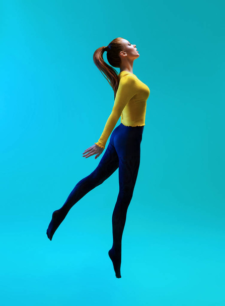 Side view of woman with ponytail wearing yellow jumper and blue leggings jumping on blue background - Foto, immagini