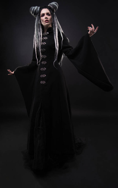 Full length view of woman with dreads wearing black gothic coat and posing on dark background - Foto, Imagen