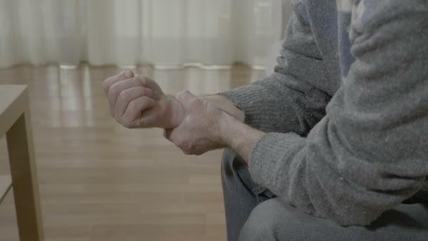 Closeup of old man with arthritis touching his painful wrist having rheumatism sitting on the couch at home - Πλάνα, βίντεο