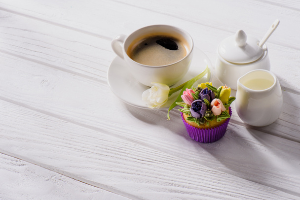 close up view of cup of coffee, tulip flower, sweet muffin and jag of cream on white wooden tabletop - Fotografie, Obrázek