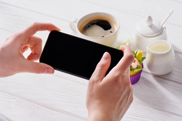 przycięte zdjęcie kobiety z smartphone rezygnować pusty ekran, filiżanka kawy, jag krem i słodki muffin na powierzchni drewnianych - Zdjęcie, obraz