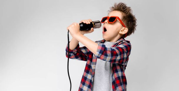 Beau garçon caucasien préscolaire en tenue décontractée posant dans des lunettes de soleil de fête avec microphone et montrant différentes expressions sur le mur blanc en studio
. - Photo, image