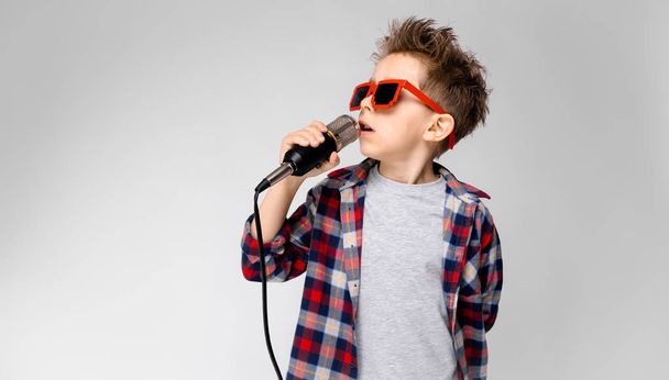 Szép óvodás kaukázusi fiú alkalmi ruhában pózol party napszemüveg, mikrofon, és bemutatja a különböző kifejezések fehér falon a stúdió. - Fotó, kép