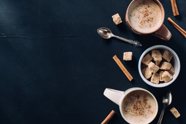 top view of cups of coffee, spoons, bowl of cane sugar and cinnamon stick on dark tabletop - Zdjęcie, obraz