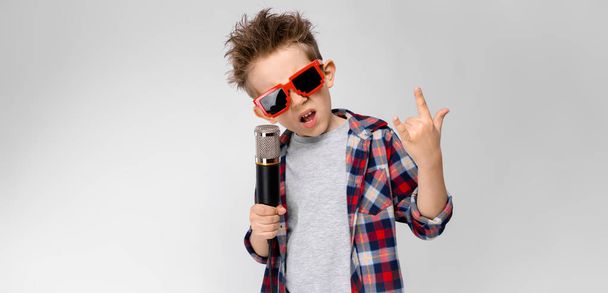 Nice preschooler caucasian boy in casual outfit posing in party sunglasses with microphone and showing different expressions on white wall in studio. - Photo, Image