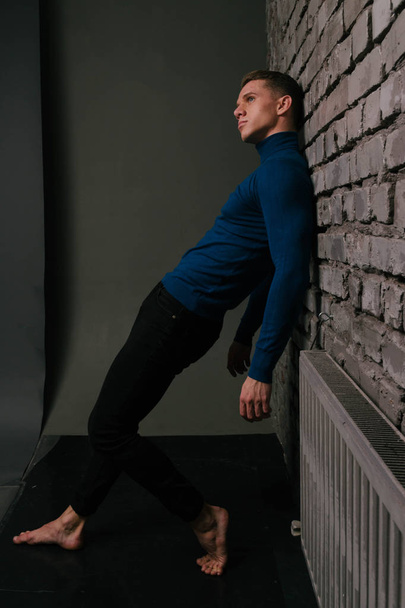 white attractive guy is nearby a brick wall background dressed in a blue sweater and black pants - Fotoğraf, Görsel