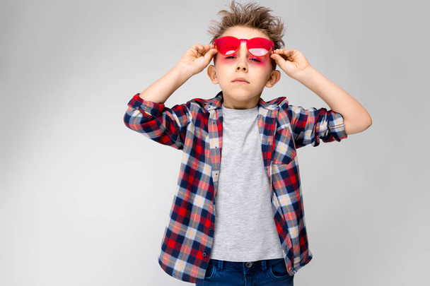 Niza niño preescolar caucásico en traje casual y gafas de sol rojas que muestran diferentes expresiones en la pared blanca en el estudio
. - Foto, imagen