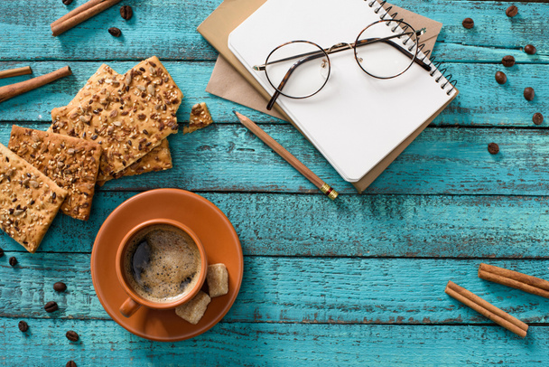 flat lay with cup of coffee, cookies, eyeglasses, empty notebook, roasted coffee beans and cinnamon sticks around on blue wooden tabletop - Photo, Image