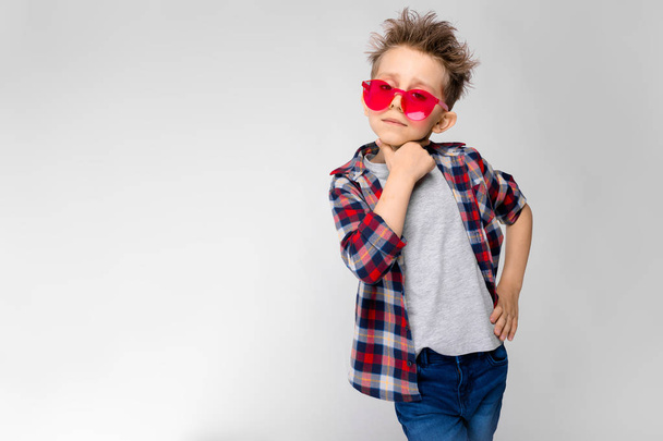 Nice caucasian preschooler boy in casual outfit and red sunglasses showing different expressions on white wall in studio. - Foto, afbeelding