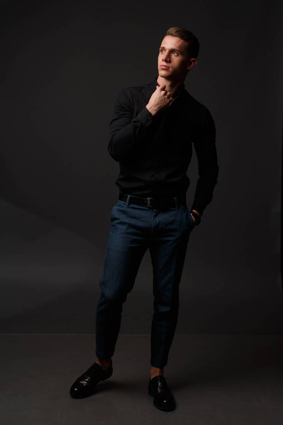 attractive white man in black shirt and blue pants stands on a dark background - Foto, Imagem