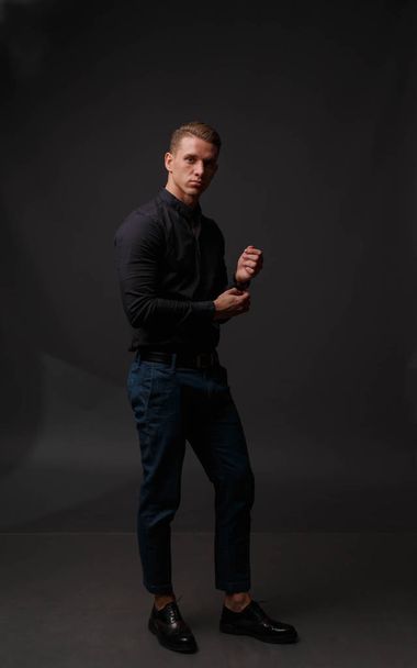 atractivo hombre blanco en camisa negra y pantalones azules se levanta sobre un fondo oscuro
 - Foto, Imagen