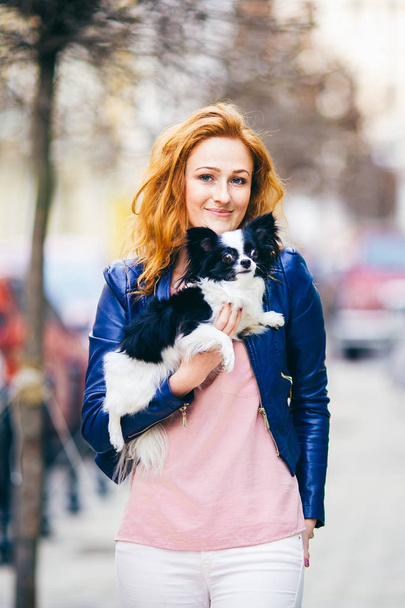 Tárgya az ember és a kutya. fiatal vörös hajú kaukázusi nő-arcán szeplőkkel rendelkezik fekete-fehér bozontos chihuahua tenyészt kutya. A lány öltözött kék bőrkabát, áll a forgalmas utcán tavasszal - Fotó, kép