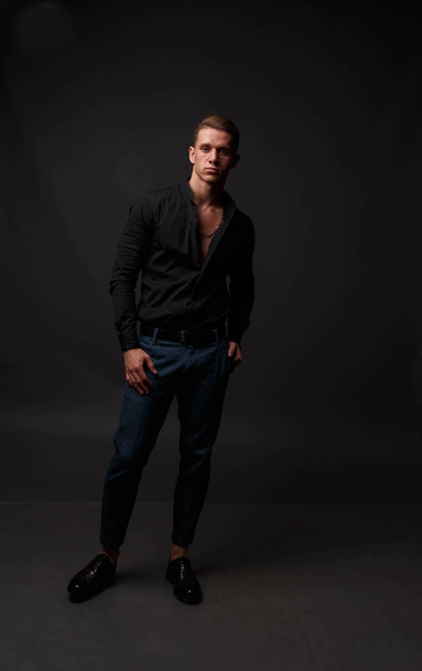 attractive white man in black shirt and blue pants stands on a dark background - Фото, изображение