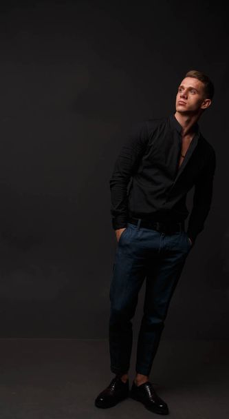 attractive white man in black shirt and blue pants stands on a dark background - Zdjęcie, obraz