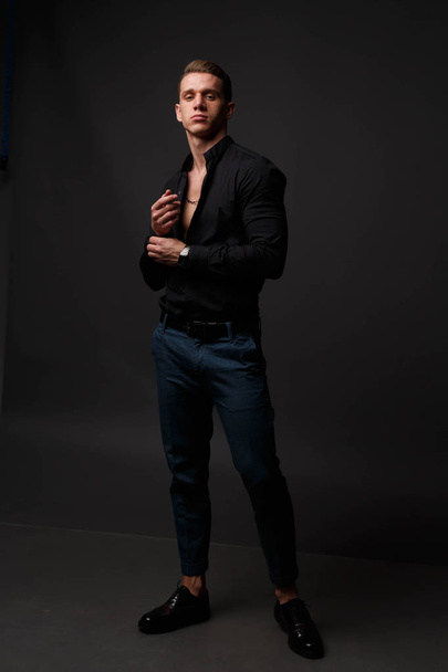 attractive white man in black shirt and blue pants stands on a dark background - Foto, Imagen