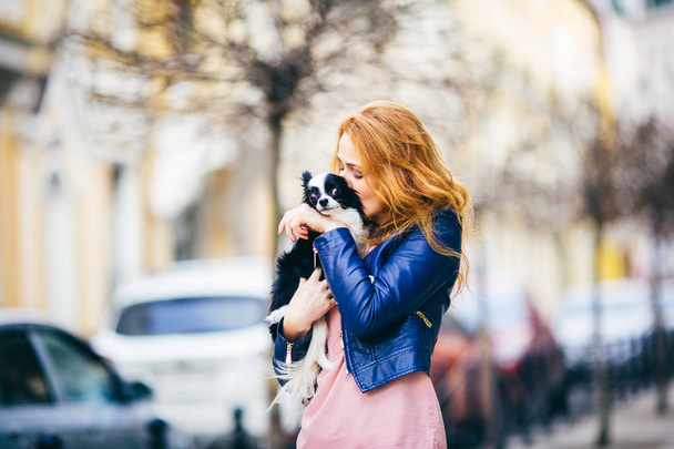 Una joven mujer caucásica pelirroja con pecas sostiene y besa, abrazando perro peludo blanco y negro de raza Chihuahua. chica vestida con chaqueta de cuero azul, se para en la calle en primavera en Europa
 - Foto, Imagen