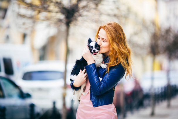 Młody redhaired kaukaski kobiety z piegami posiada i całuje, obejmując biało-czarny kudłaty pies rasy Chihuahua. Dziewczyna ubrana w niebieski skórzana kurtka, stoi na ulicy na wiosnę w Europie - Zdjęcie, obraz