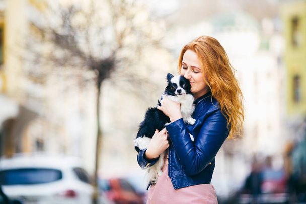 Egy fiatal redhaired kaukázusi nő szeplő tartja, és megcsókolja, átfogó fekete-fehér bozontos kutya-Chihuahua fajta. kék bőrkabát, öltözött lány áll street tavasszal Európában - Fotó, kép