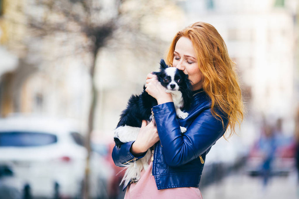 Egy fiatal redhaired kaukázusi nő szeplő tartja, és megcsókolja, átfogó fekete-fehér bozontos kutya-Chihuahua fajta. kék bőrkabát, öltözött lány áll street tavasszal Európában - Fotó, kép