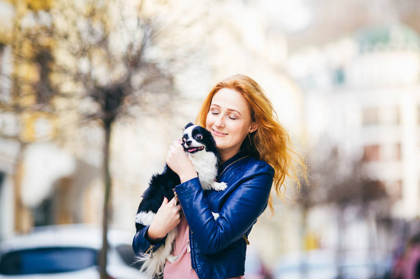 Молодая рыжая белая женщина с веснушками на лице держит и целует, обнимает, любит черно-белую мохнатую собаку чихуахуа. Девушка в синей кожаной куртке, стоящая на улице весной
 - Фото, изображение