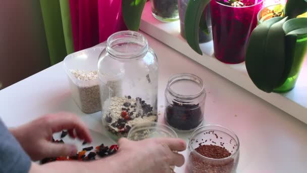 The man mixes in a glass jar the dried fruits, oat flakes and flax seed with coconut shavings for muesli. - Imágenes, Vídeo
