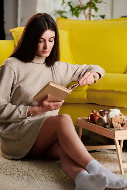 Nainen lukee kirjaa istuessaan lattialla lähellä puinen tarjotin kuppi kahvia ja leivonnaisia kotona
 - Valokuva, kuva