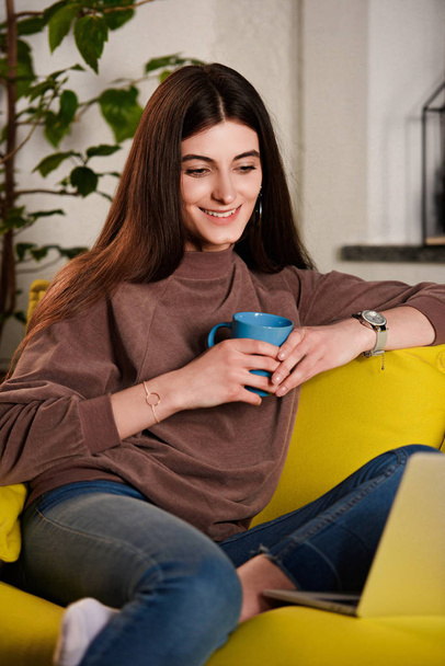 mujer con taza de café usando el ordenador portátil en el sofá en casa
 - Foto, Imagen