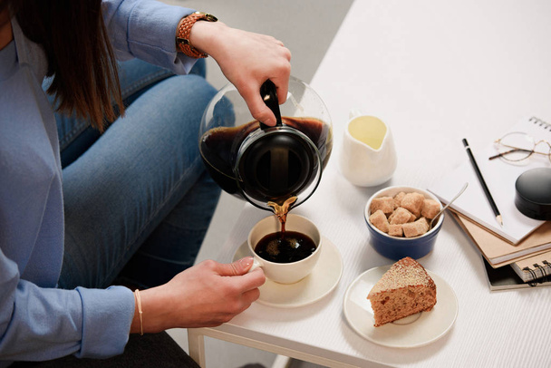 tiro recortado de la mujer vertiendo café en la taza con azúcar de caña y pedazo de pastel en la mesa de café
 - Foto, Imagen